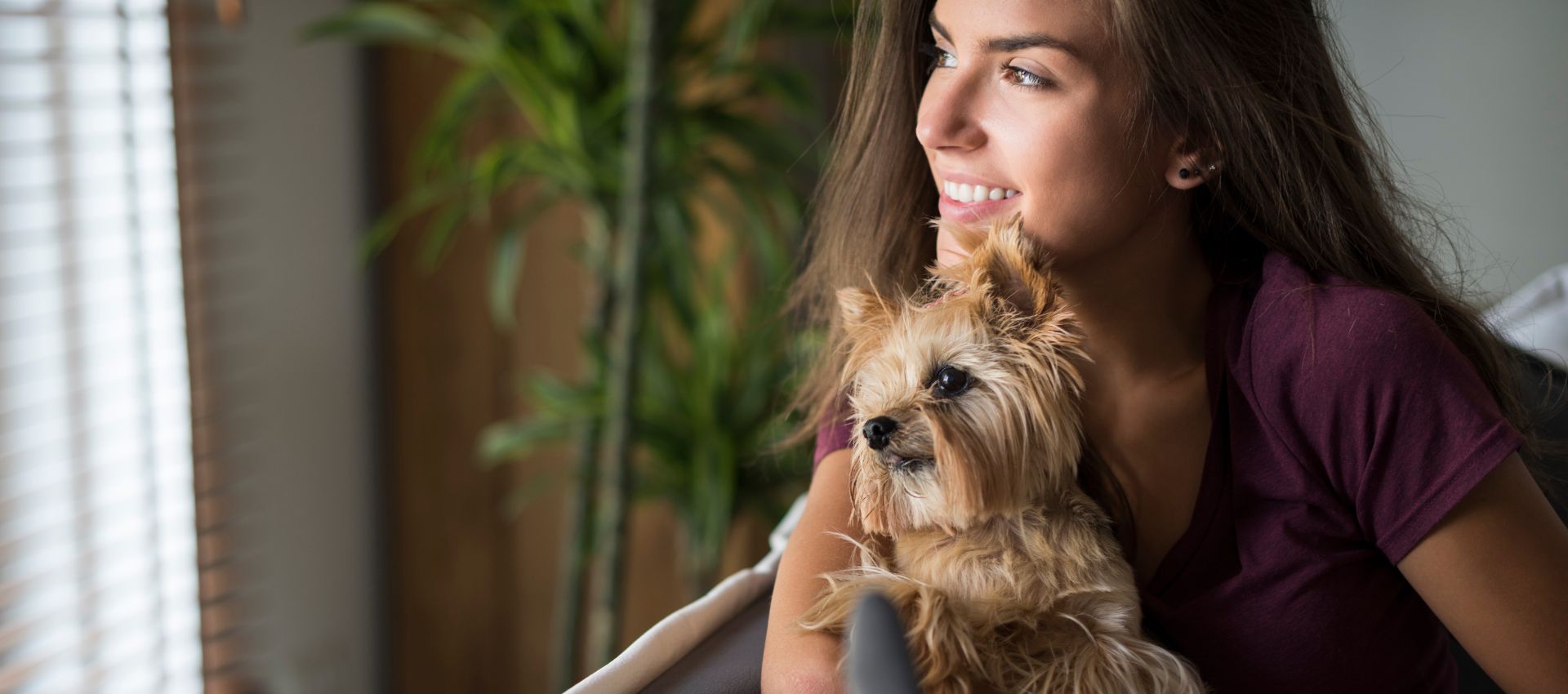 Mujer sonriendo junto a su perro de raza pequeña mirando por la ventana de su condominio en Miami.